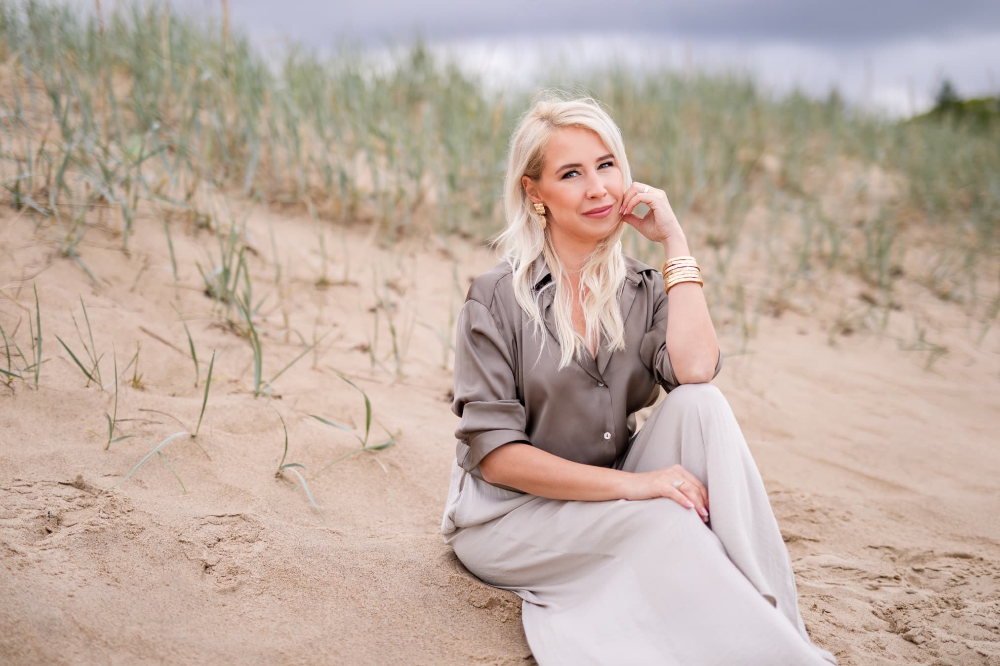 Svetlana Ambre дружелюбно улыбается, сидя на песке с травой на пляже