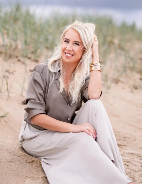 Svetlana Ambre дружелюбно улыбается, сидя на песке с травой на пляже