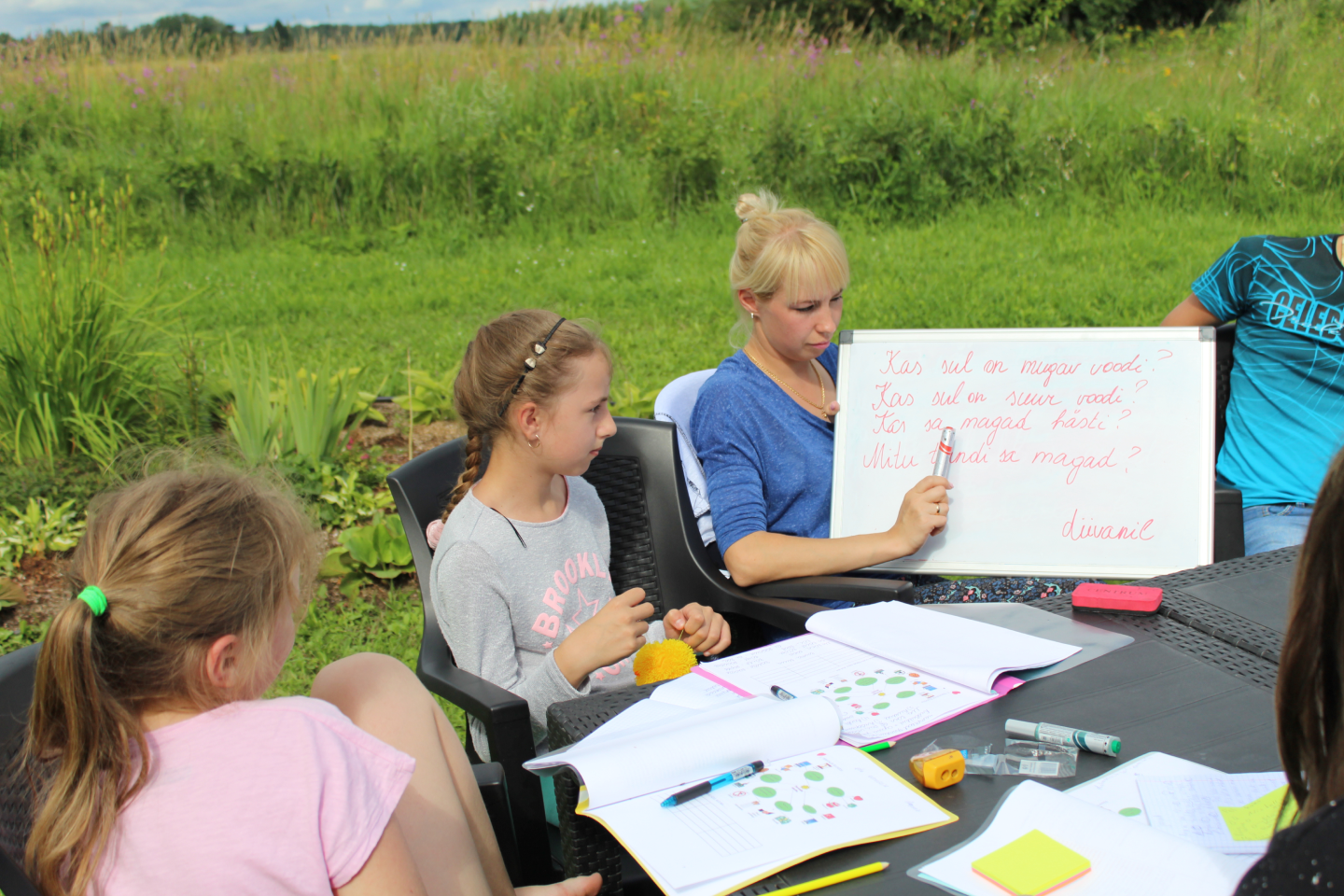 На фоне зеленого поля и леса дети учатся у Svetlana Ambre и смотрят на плакат в ее руках